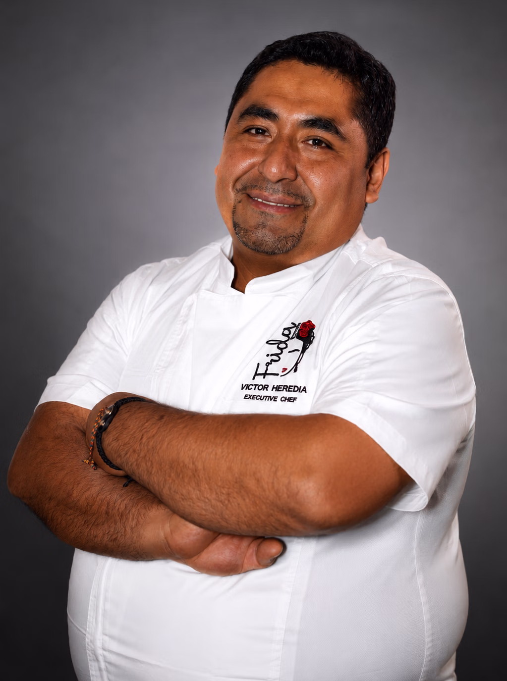 Portrait of Victor Heredia, executive chef, smiling with arms crossed in a white chef coat labeled Victor Heredia Executive Chef
