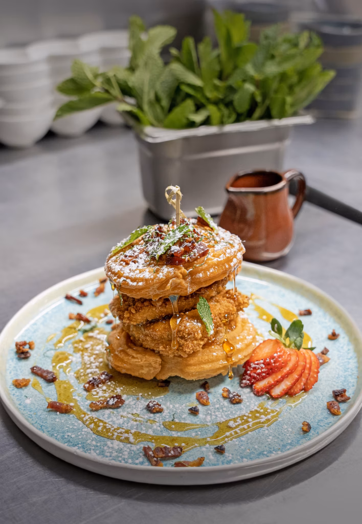 Stack of fried chicken and waffles on a blue plate, drizzled with syrup and dusted with powdered sugar, garnished with mint and strawberries in the foreground.